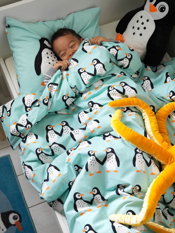 Chlapec zaspáva v posteli v svetle tyrkysovej posteľnej bielizni s tučniakmi BLÅVINGAD, vedľa neho sú plyšové hračky, tučniak a chobotnica BLÅVINGAD.