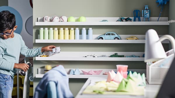Chłopiec porządkujący piłki, butelki, zabawki, papierowe modele i samochodziki na czterech białych półkach na zdjęcia MOSSLANDY zamontowanych na ścianie.