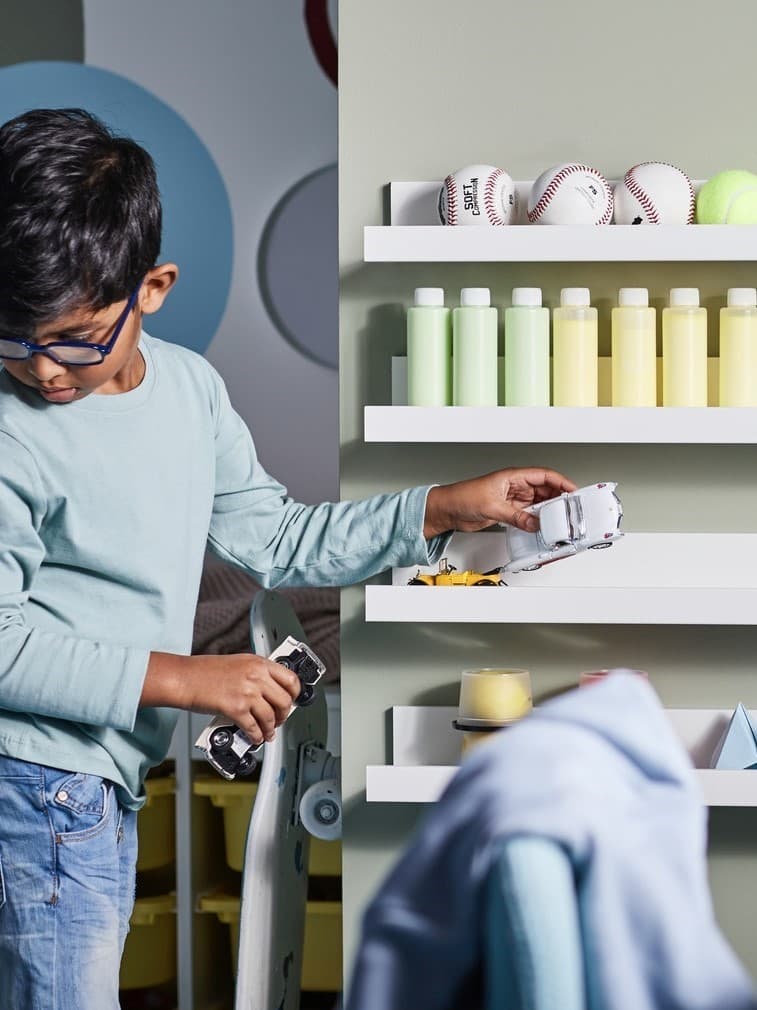 Chłopiec porządkujący piłki, butelki, zabawki, papierowe modele i samochodziki na czterech białych półkach na zdjęcia MOSSLANDY zamontowanych na ścianie.