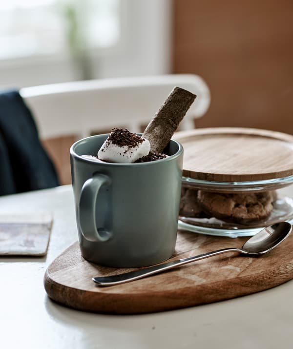 Chocolat chaud et shamallows dans un mug gris-bleu posé sur une planche à découper avec des biscuits dans un bocal en verre avec un couvercle en bambou.