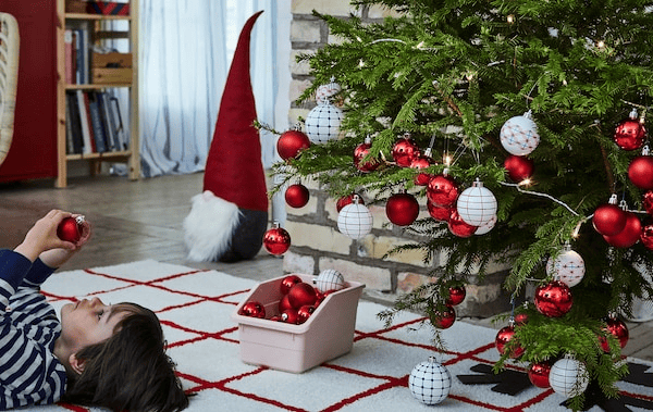 Christmas tree heavy with decorations with a young boy lying face up on rug next to it, inspecting an ornament.