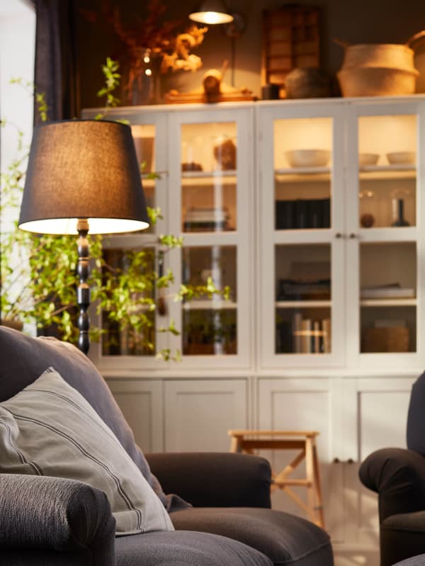Čierna stojacia lampa KINNAHULT stojí pri tmavosivej pohovke ESSEBODA, v pozadí je biela úložná zostava HAVSTA.