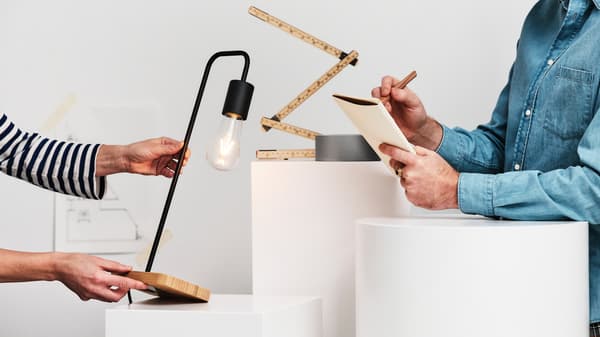 Cilvēks zilā džinsa kreklā raksta piezīmju grāmatiņā, kamēr cits cilvēks pasniedz melnu/bambusa TVÄRHAND galda lampu.
