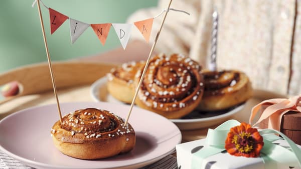 Cinnamon bun with a festive banner on a pink plate, surrounded by pastries, gifts, and a birthday candle.