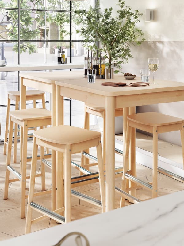 Cinq tabourets de bar RÖNNINGE autour de deux tables de bar sur lesquelles se trouvent des verres de vin et d’eau, près d’une fenêtre.