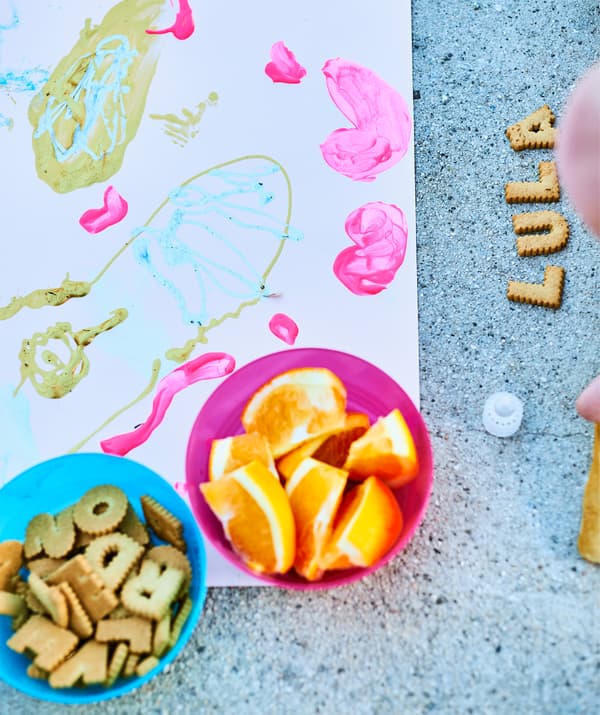 Ciotole con biscotti e spicchi d'arancia, il disegno di un bambino e biscotti a forma di lettere che formano il nome LULA su un pavimento in cemento - IKEA