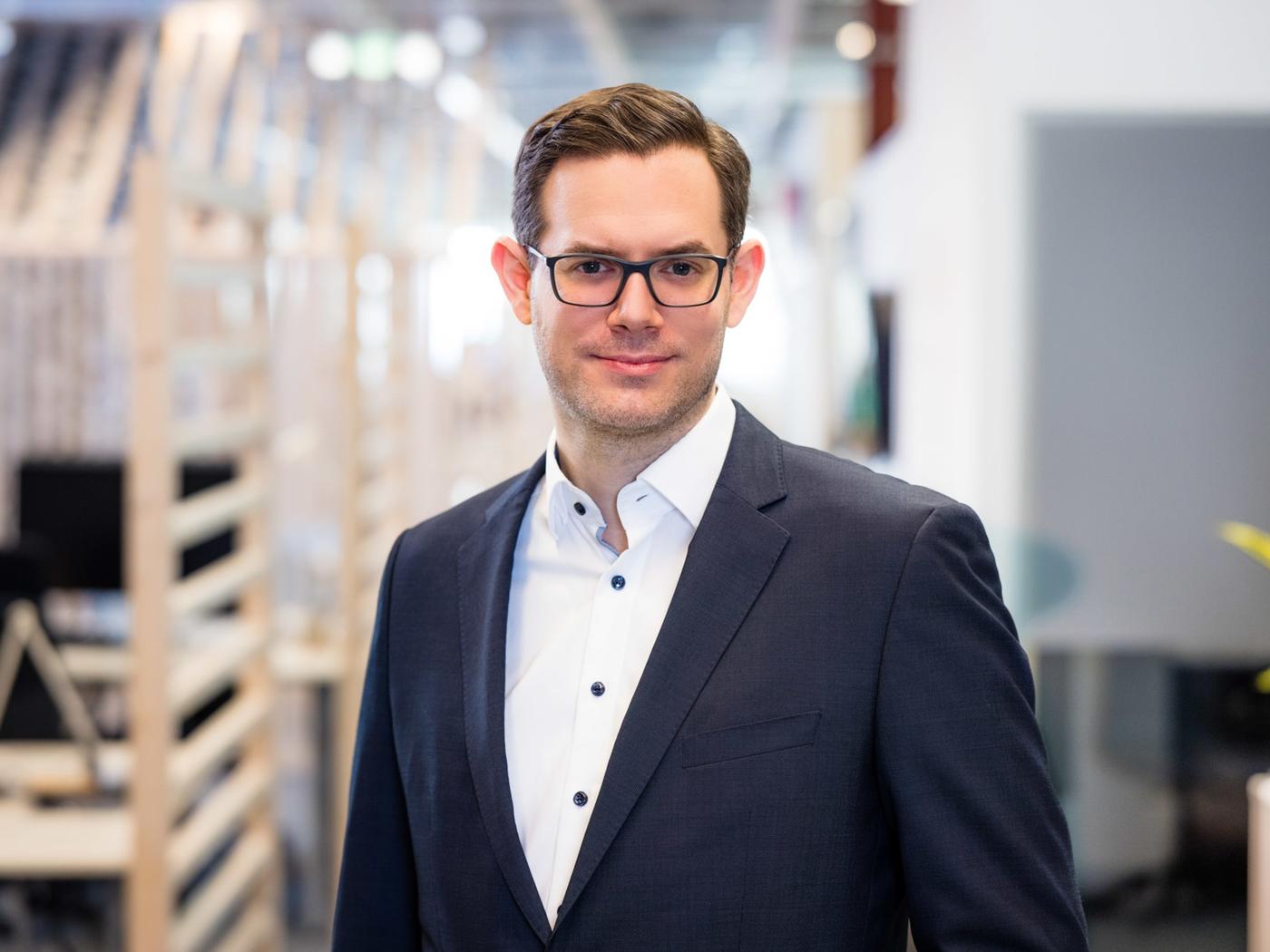 Claudio Winkler in einem Anzug steht lächeldn in einem Büro