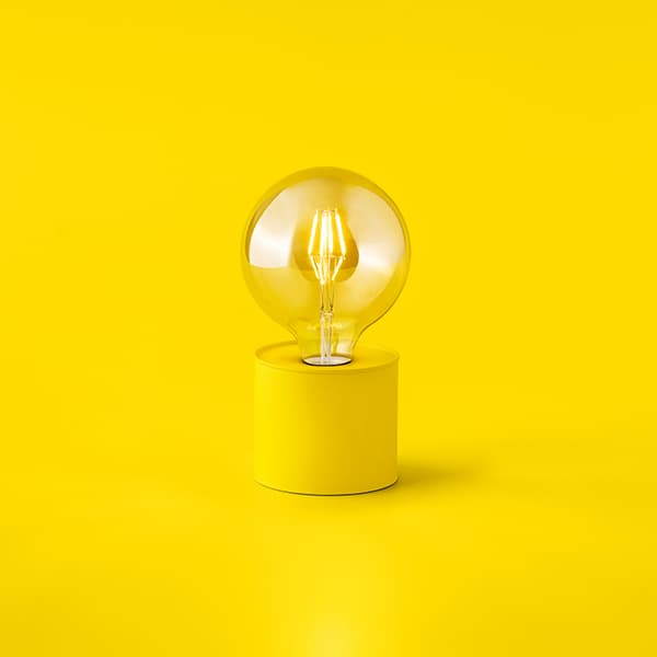 Clear glass light bulb with visible filament mounted on a yellow cylindrical base, set against a bright yellow background.