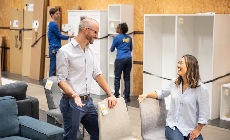Clienți într-un magazin IKEA discută și testează scaune în zona de mobilier, în timp ce angajați în uniforme albastre „Hej!” lucrează la montarea și organizarea dulapurilor în fundal.