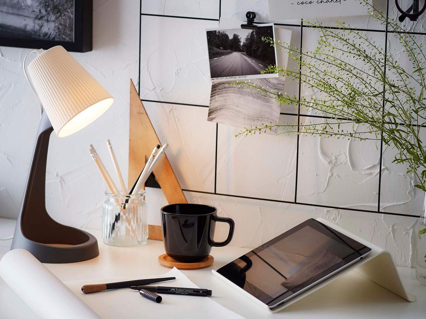 Close up image of a white desk top with a black wire notice board above with various papers and items hanging on it. The desk had a work lamp, mug, pencil cup and tablet on it.