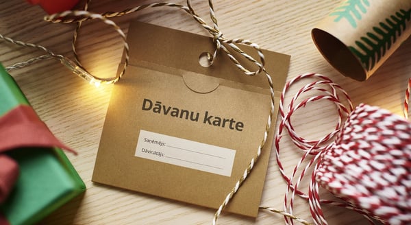 Close-up of a brown IKEA gift card envelope with Latvian text 'Dāvanu karte' surrounded by Christmas wrapping items, twine, and festive lights.