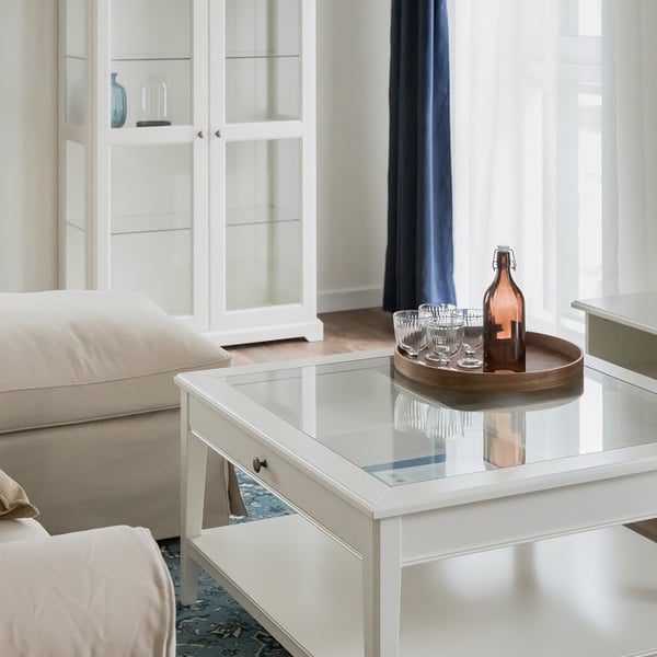 Close-up of a classic white glass-top coffee table in a bright living room. A round wooden tray on the table holds an amber glass bottle and several small drinking glasses. Behind the table, a beige sofa and a tall white display cabinet with glass doors are visible. The room features dark navy curtains, sheer white drapes, and a patterned blue area rug, creating a sophisticated and airy atmosphere.