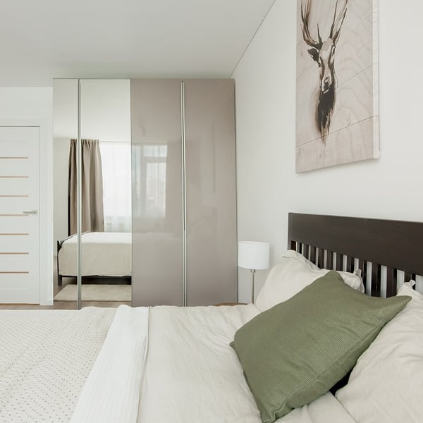 Close-up of a cozy bedroom corner with a bed dressed in neutral bedding and a large green pillow. Above the dark wooden headboard is a canvas print featuring a deer. A modern wardrobe with high-gloss taupe and mirrored panels reflects the natural light coming through sheer curtains, enhancing the bright and clean atmosphere.