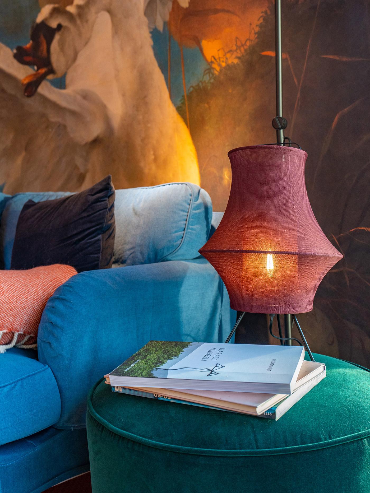 Close-up of a cylindrical side table made of fabric with books on it and a lamp with a fabric lampshade hanging above it