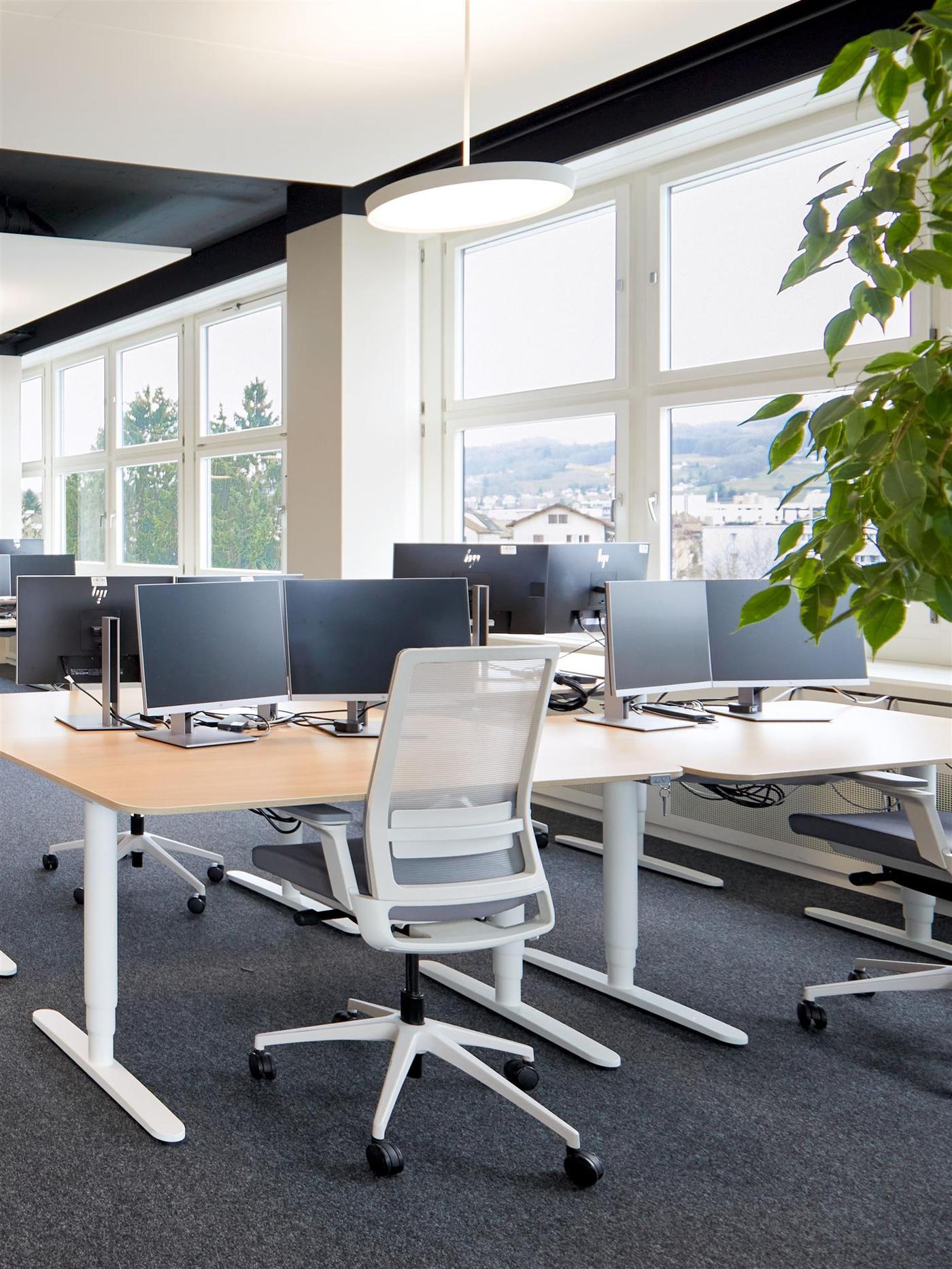 Close-up of a desk with a work chair and monitors in an open-plan office by the window