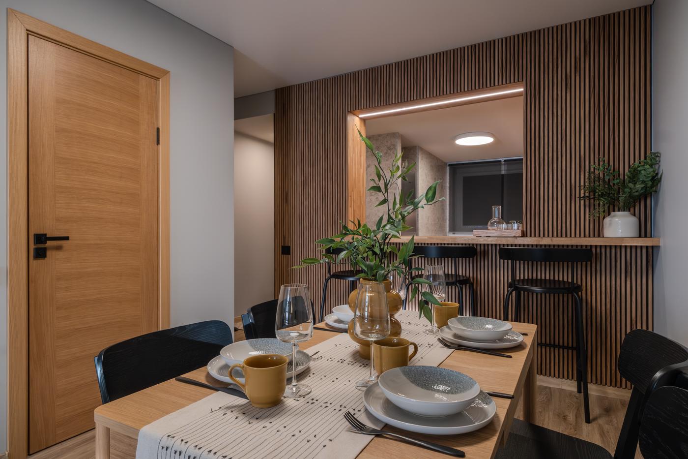 Close-up of a dining table set with plates, glasses and mugs. A wooden kitchen bar with stools is visible in the background.