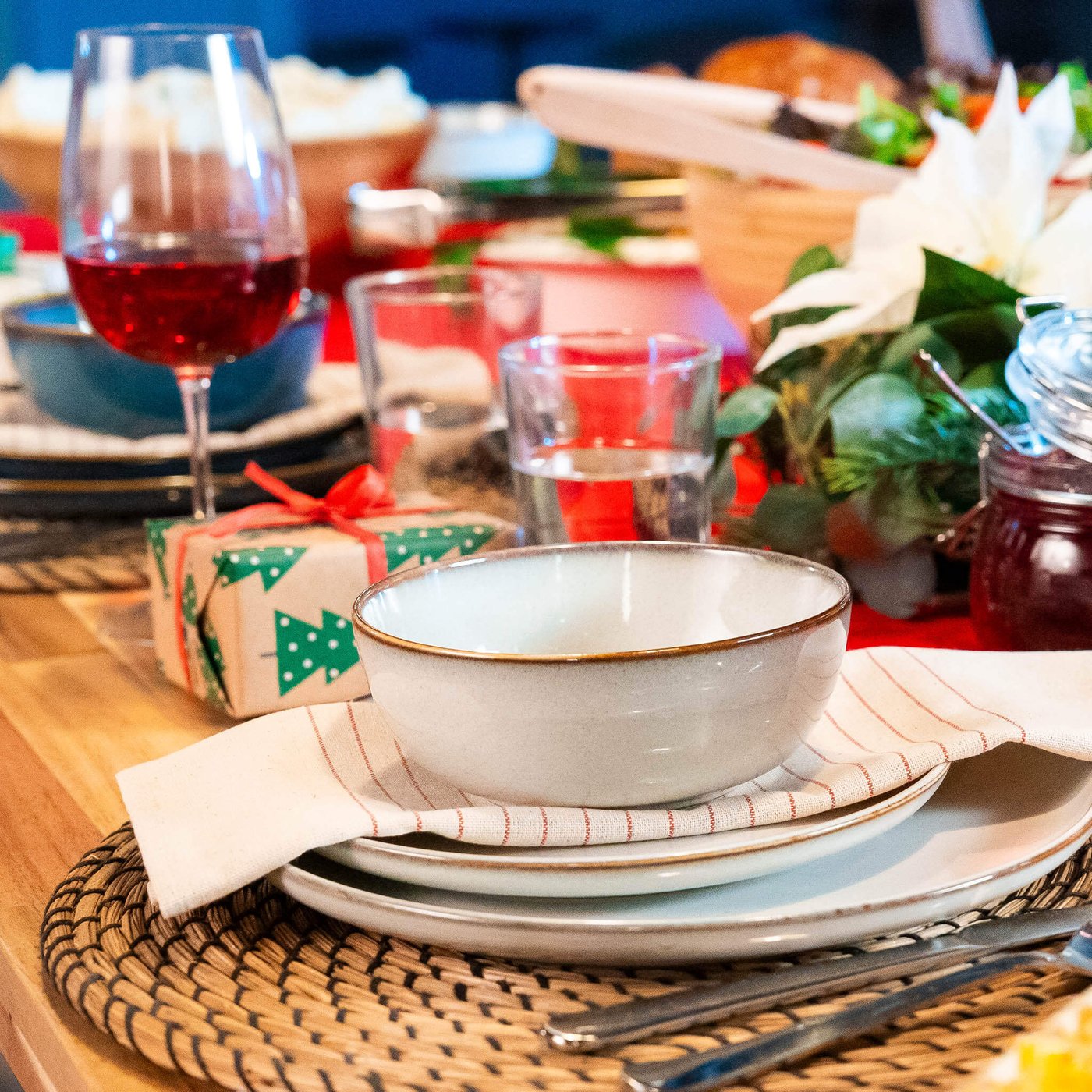 Close-up of a holiday table setting with a white bowl on a plate, wine glass, wrapped gift, and festive greenery centerpiece.