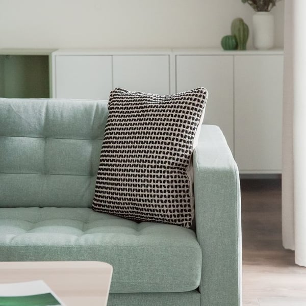 Close-up of a light mint green tufted sofa with a decorative black and white patterned throw pillow. In the background, a minimalist white sideboard with clean lines stands against the wall, topped with small green ceramic decor and a white vase holding foliage. The room features a soft and modern color palette with natural wood flooring and neutral curtains, creating a cozy and contemporary atmosphere.