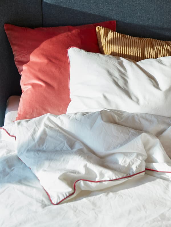 Close-up of a made bed with red, white, and light blue cushions and throws in soft natural lighting.