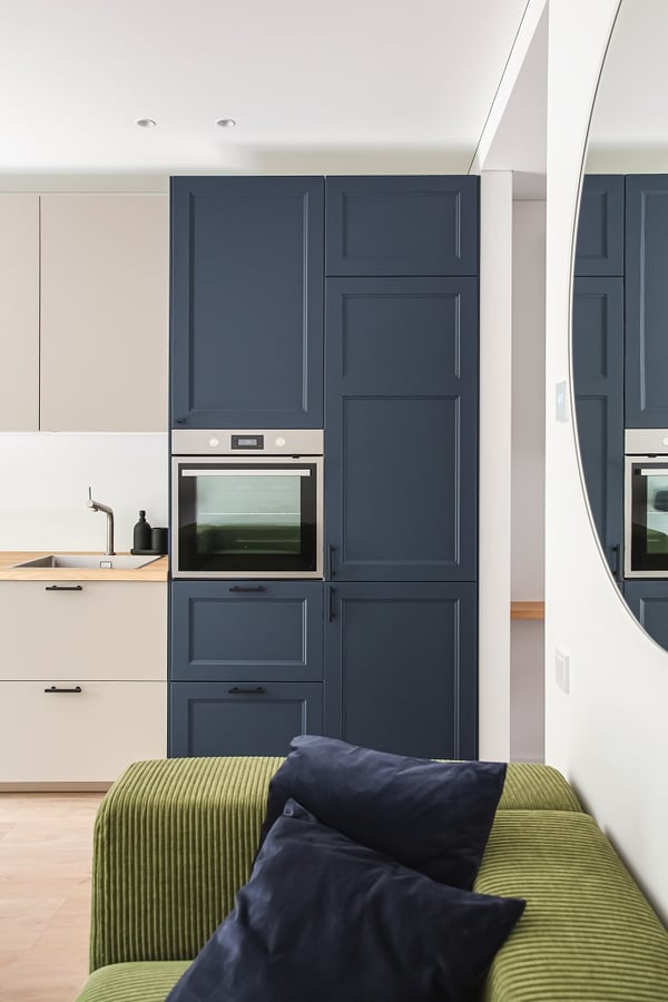 Close-up of a modern kitchen corner featuring deep navy blue cabinetry with integrated oven and tall storage units. Light beige lower cabinets with a wood countertop and matte black hardware contrast elegantly with the darker elements. Part of a green corduroy sofa with navy throw pillows is visible in the foreground, alongside the edge of a round wall mirror, reflecting the cohesive and stylish design of the living space.