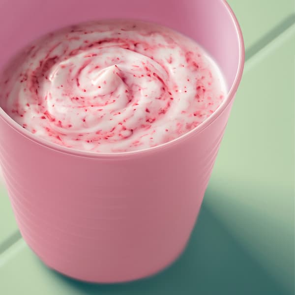 Close-up of a pink kid’s KALAS mug filled with creamy pink smoothe, shown on a light green table outdoors.