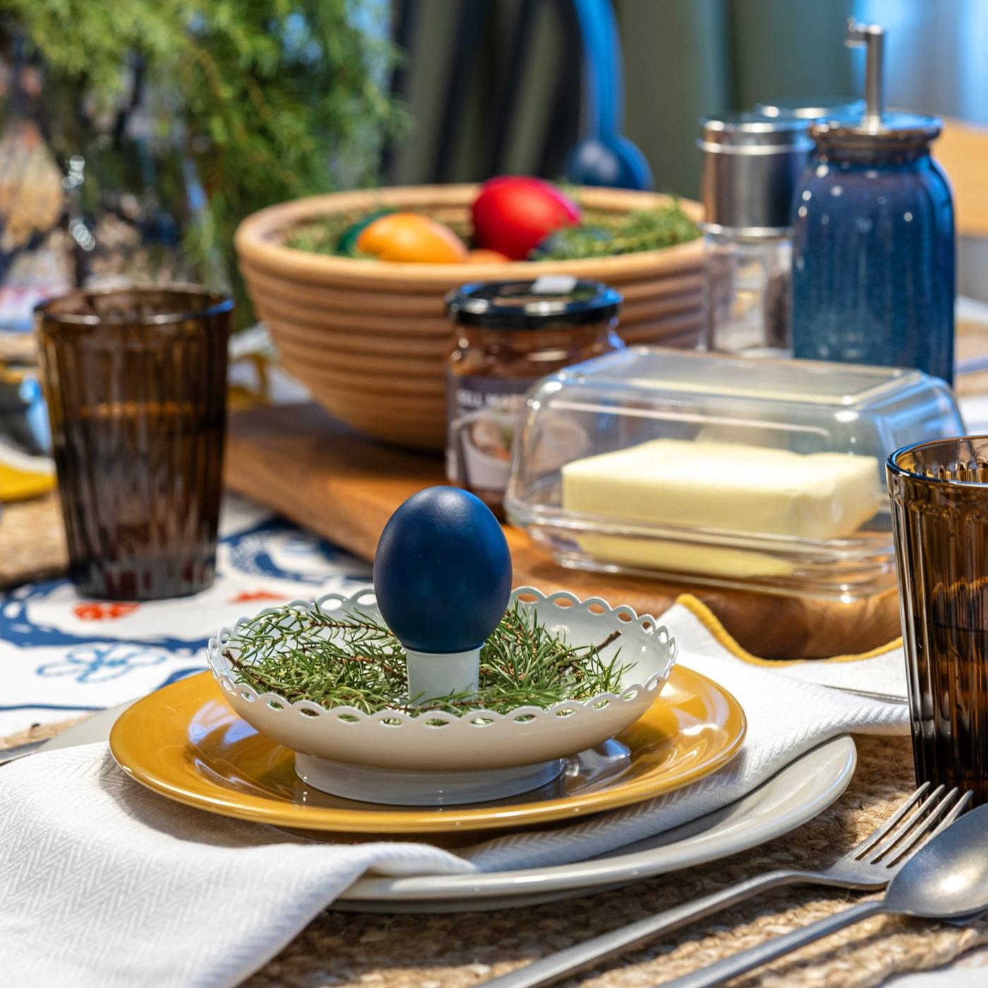 Close-up of a place setting with layered plates, a yellow napkin, woven placemat and a painted egg placed on top of a candlestick.