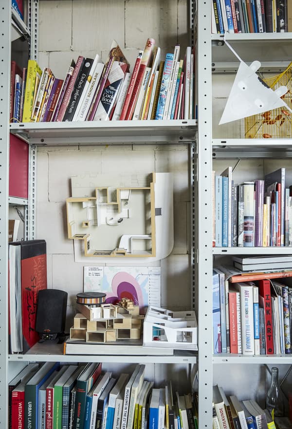 Close-up of an adjustable open storage unit in gray metal stuffed books and some 3D cardboard house prototypes.