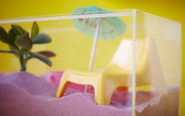 Close-up of an IKEA BJÖRNARP acrylic display box filled with pink sand, a doll’s easy chair, a potted plant and a drink umbrella.