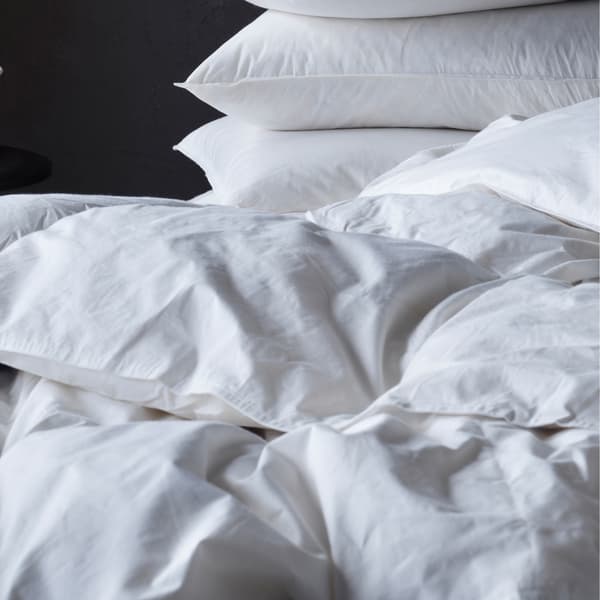 Close-up of an untidy crisp white duvet on top of a bed, with a neat pile of white pillows behind it.