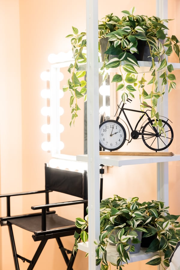 Close-up of decorative shelf with potted plants, bicycle-shaped clock, and makeup chair in background.