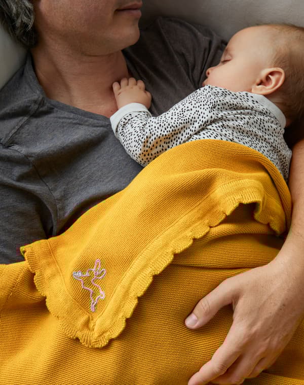 Close-up of father lying with baby on his chest with arm around the baby and SOLGUL yellow blanket over them.