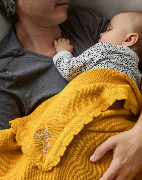 Close-up of father lying with baby on his chest with arm around the baby and SOLGUL yellow blanket over them.
