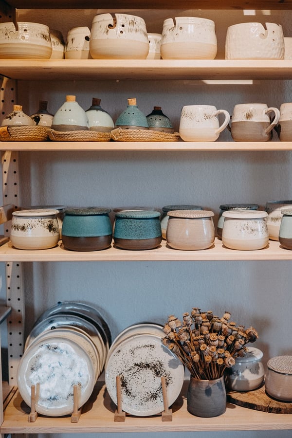 Close-up of handmade ceramic cups, bowls, and plates on natural wood shelves.