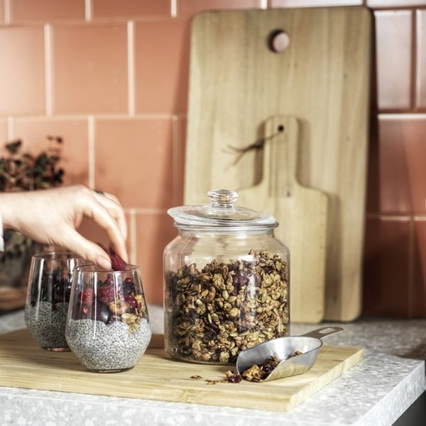 Close-up of hands taking granola from a VARDAGEN glass jar and sprinkling it onto chia pudding on a kitchen worktop.