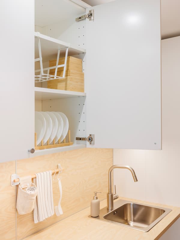 Close-up view of a kitchen featuring an open cabinet with shelf inserts for plates, a wooden wall panel, wooden countertop and a sink.