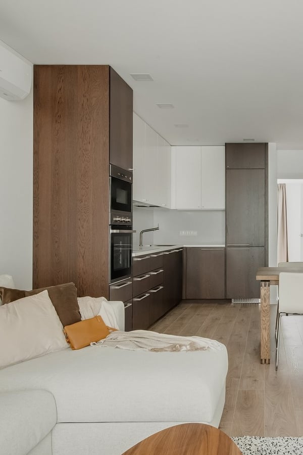 Close-up view of a modern open-plan kitchen and living area. The kitchen features a mix of dark wood and white cabinetry with sleek handles, a built-in oven, and a minimalist faucet. The living area includes a soft beige sectional sofa adorned with neutral pillows, and a wooden coffee table on a light wood floor, creating a warm and cohesive interior.
