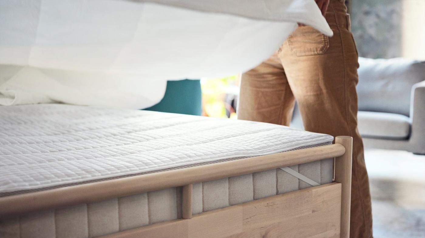 Človek naťahuje prestieradlo na matrac na drevenom ráme postele.