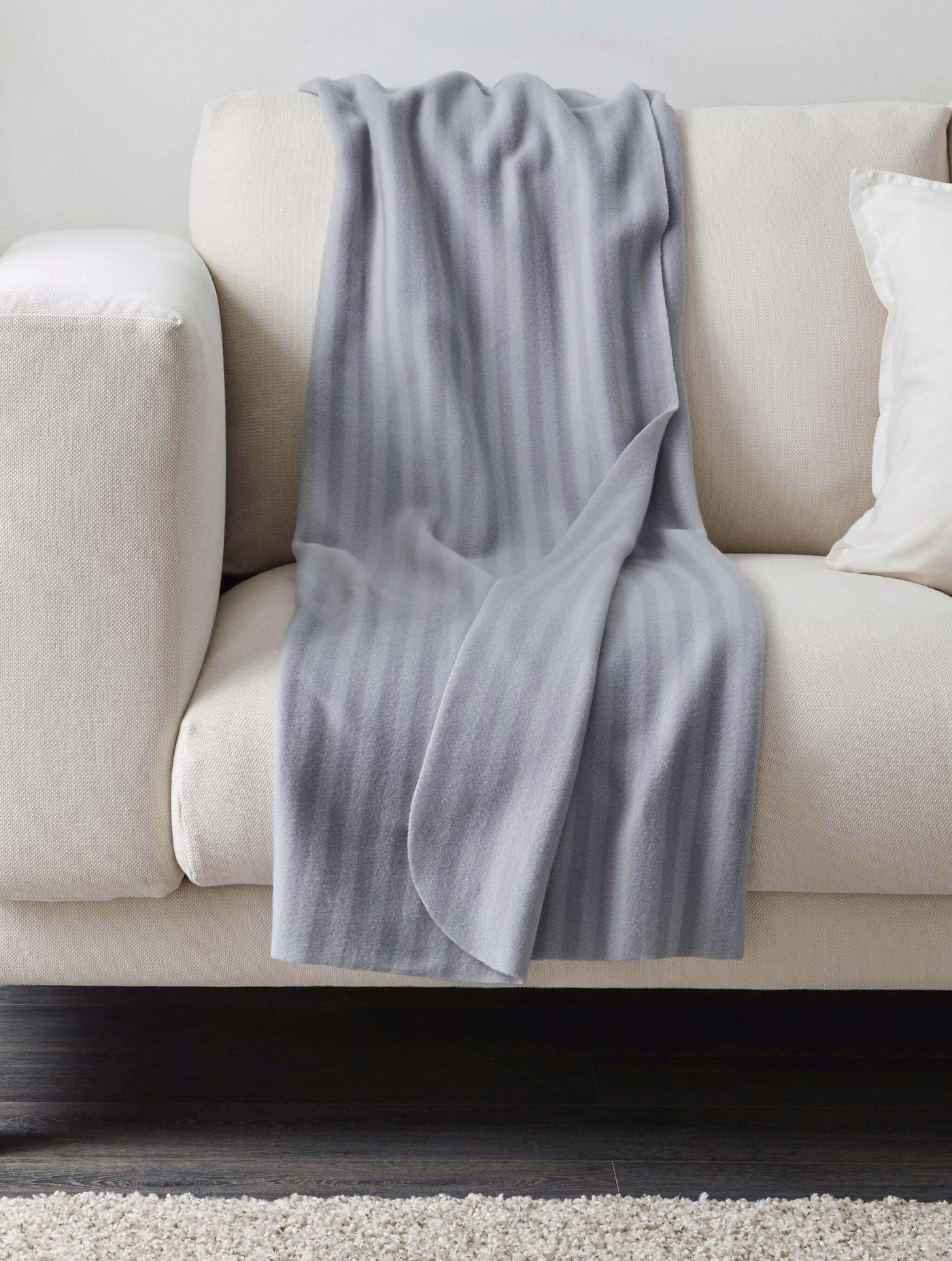 Cobija gris, posiblemente VITMOSSA de IKEA, colocada de manera casual sobre un sofá blanco. Las arrugas insinúan que alguien estuvo recostado recientemente.