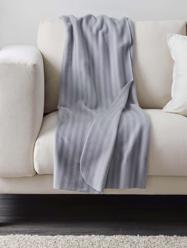 Cobija gris, posiblemente VITMOSSA de IKEA, colocada de manera casual sobre un sofá blanco. Las arrugas insinúan que alguien estuvo recostado recientemente.
