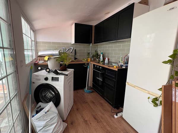 Cocina antes de ser reformada con armarios y encimera de madera, lavadora en medio, frigorífico y utensilios de cocina.