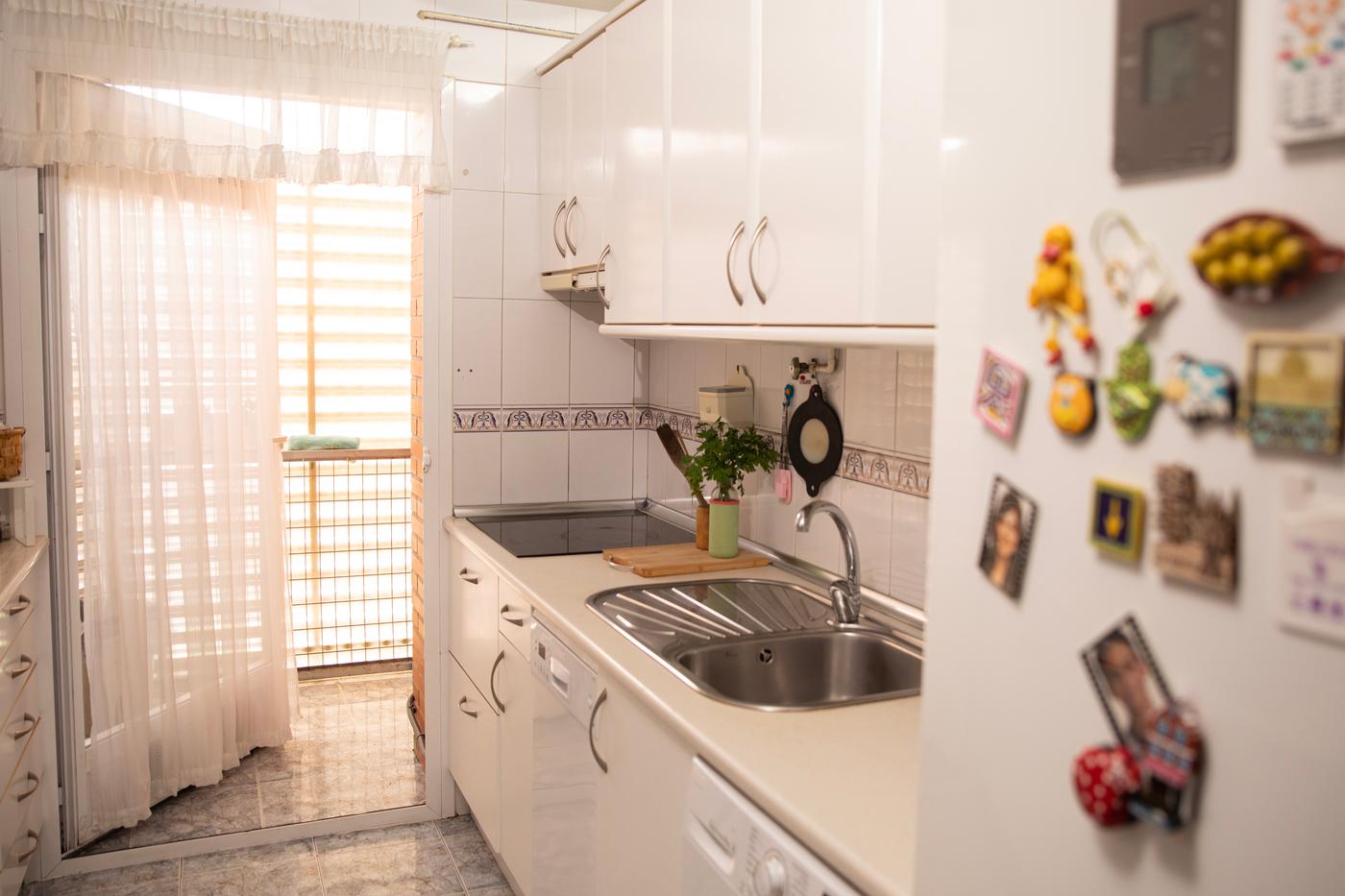 Cocina antes de ser reformada con nevera, encimera, estantería, vitrocerámica, fregadero y utensilios de cocina.