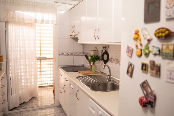 Cocina antes de ser reformada con nevera, horno, encimera, estantería, vitrocerámica, fregadero y utensilios de cocina.