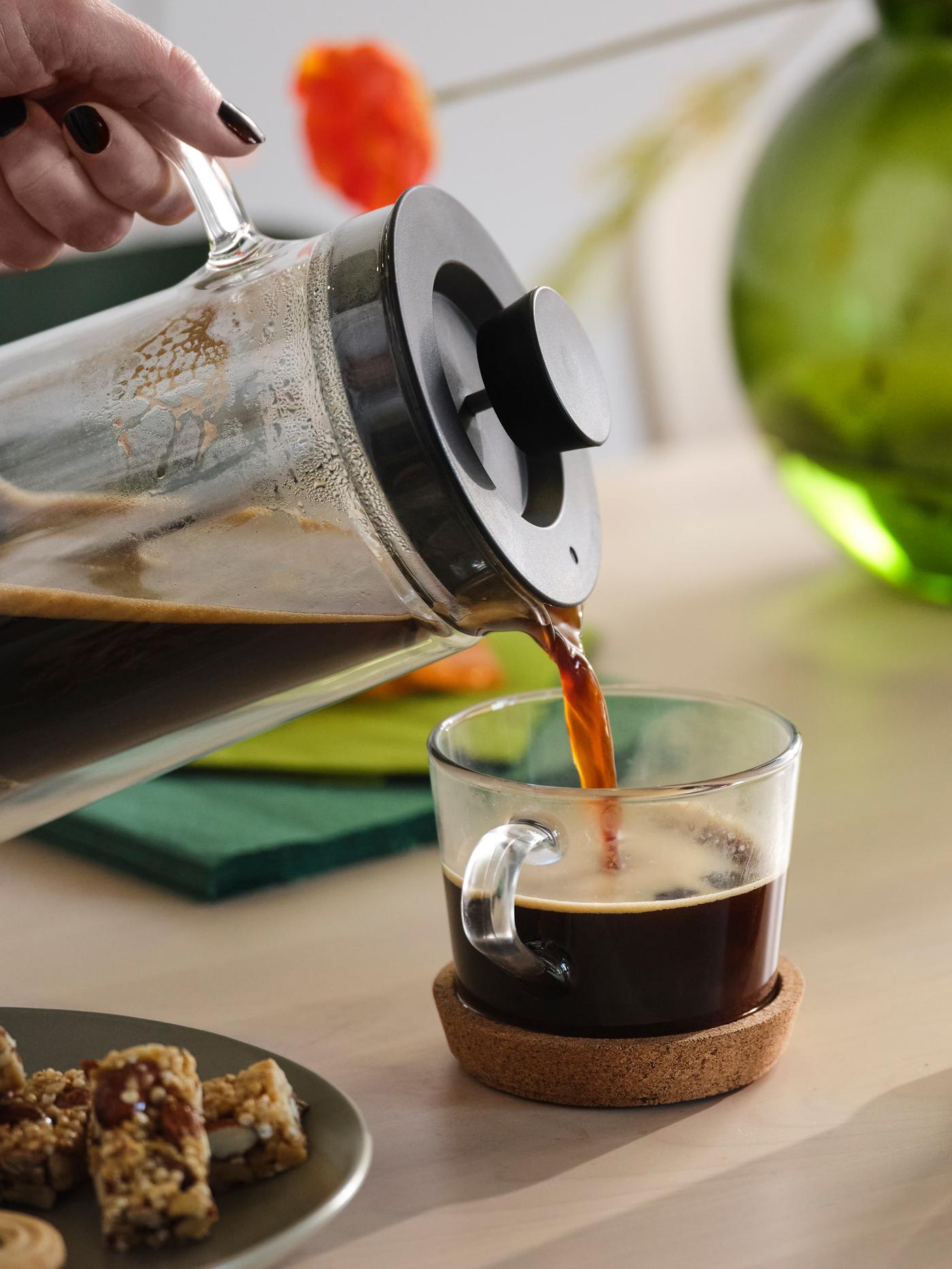 Coffee being poured from a coffee pot into a mug on a cork coaster. In the background is a green vase and orange flower.