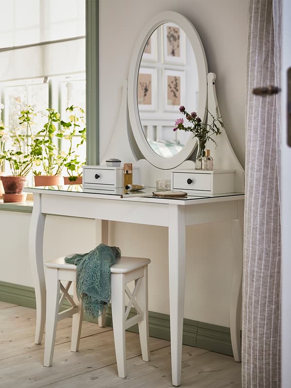 Coiffeuse blanche HEMNES avec plusieurs accessoires de beauté et un tabouret en dessous blanc , le tout dans une pièce assez lumineuse avec une fenêtre sur laquelle se trouve des pots à plantes sur le rebord