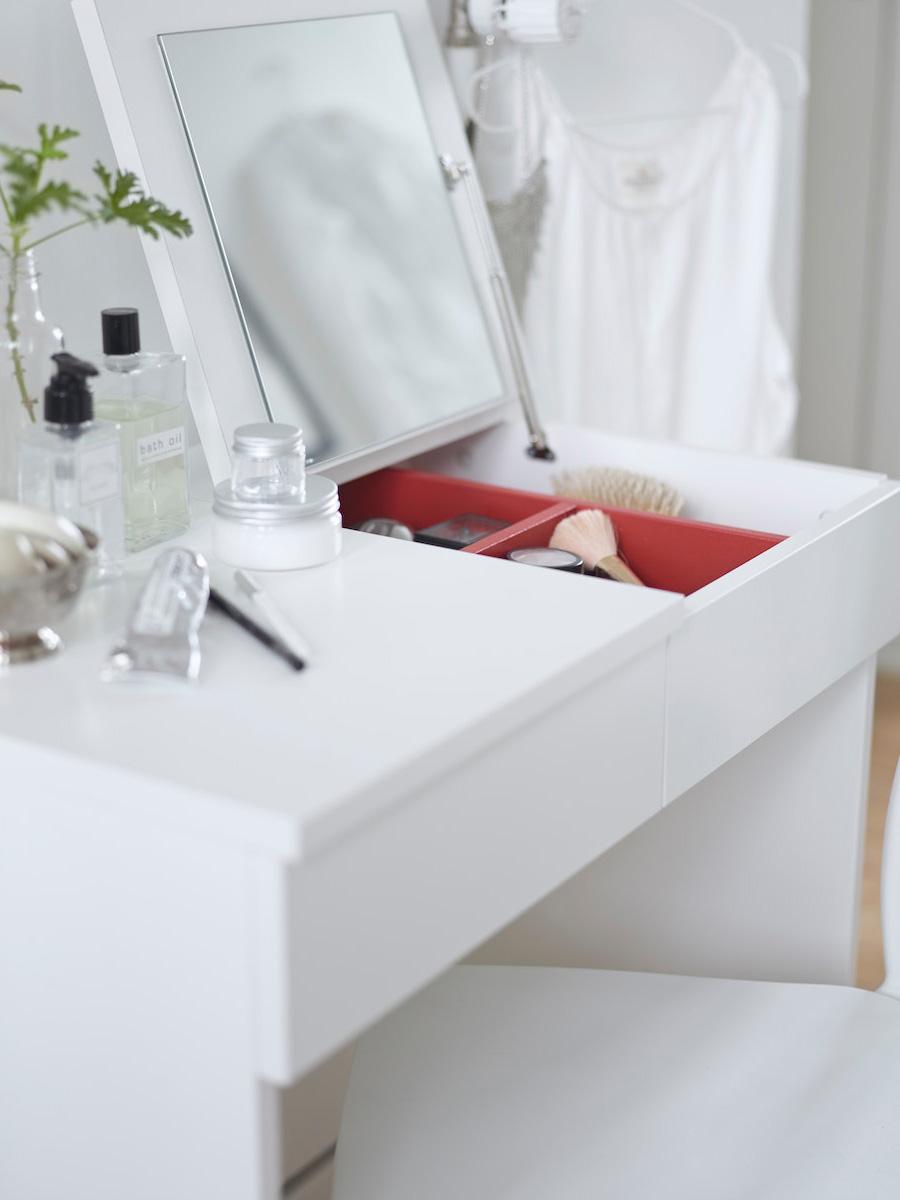 Coiffeuse BRIMNES blanche ouvert avec un miroir intégré avec à l'intérieur des compartiments contenant des pinceaux et autres accessoires de maquillage. Sur le côté fermé se trouve plusieurs pot de crème et bouteille de parfum et autres cosmétiques