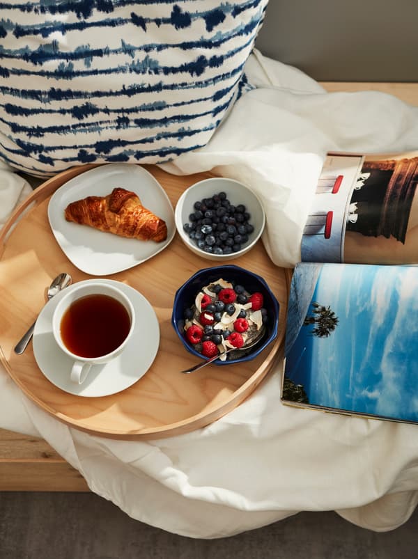 Coin de balcon où un plateau rond est posé sur du linge de lit. Sur le plateau se trouvent un bol STRIMMIG contenant du porridge aux baies et une tasse de thé.