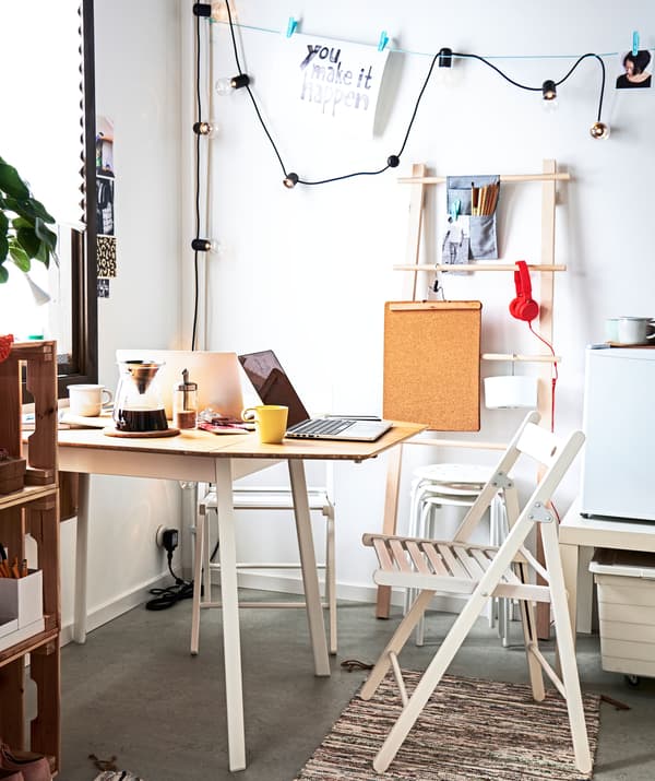 Coin de l’appartement équipé d’une table appuyée contre un mur, de chaises pliantes, de tabourets et de rangements variés.