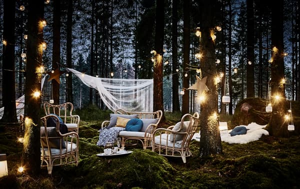 Coin détente le soir, lors d’une fête de noces organisée en forêt, avec fauteuils en rotin, nombreux coussins, guirlande lumineuse et lanternes suspendues