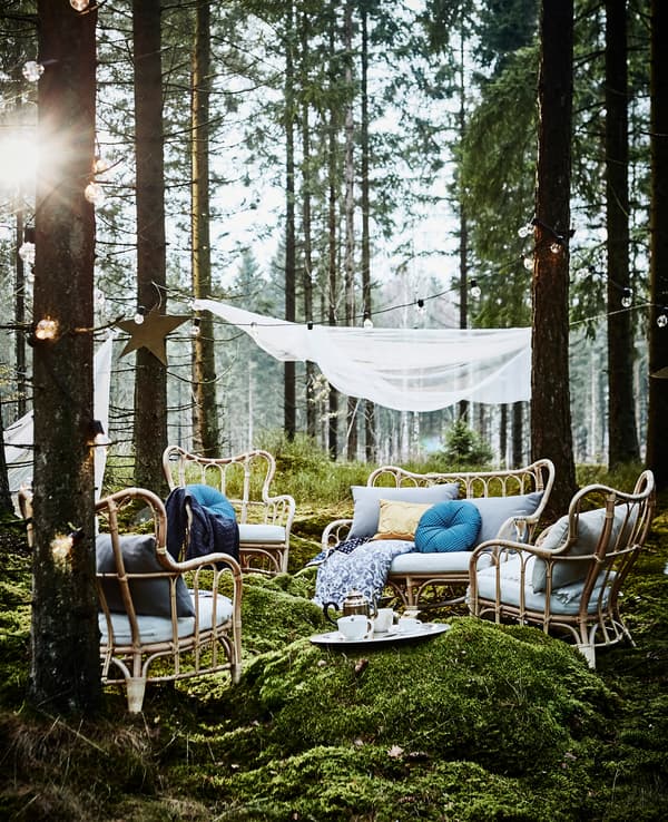 Coin détente lors d’une fête de noces organisée en forêt, avec fauteuils en rotin, nombreux coussins et guirlandes lumineuses, à côté d’un photomaton maison