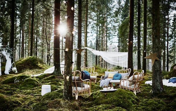 Coin détente lors d’une fête de noces organisée en forêt, avec fauteuils en rotin, nombreux coussins et guirlandes lumineuses, à côté d’un photomaton maison